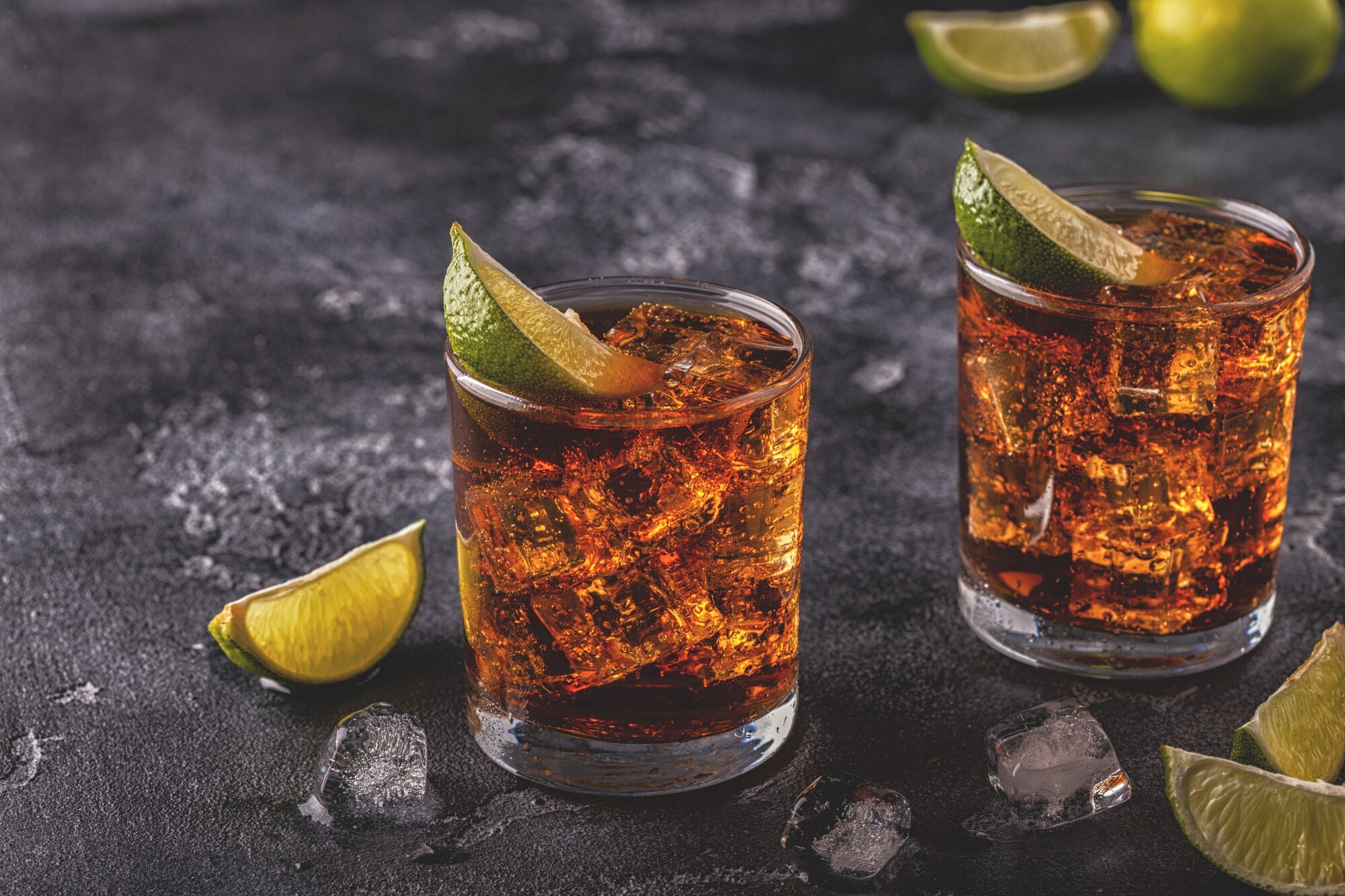 Two glasses of dark cola with ice and lime wedges on a dark stone surface.
