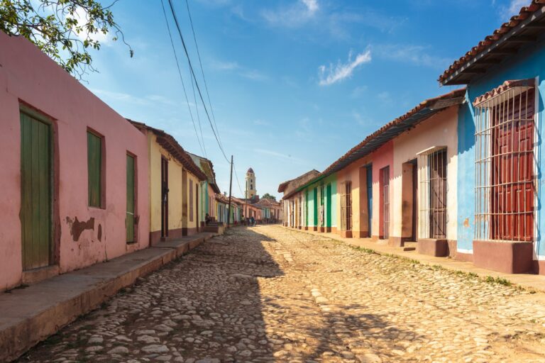Pastel houses with tiled roofs line a cobblestone street in Trinidad, Cuba, with a bell tower in the distance.