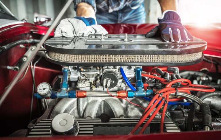 A mechanic wearing gloves leans over the exposed engine of a red classic car.