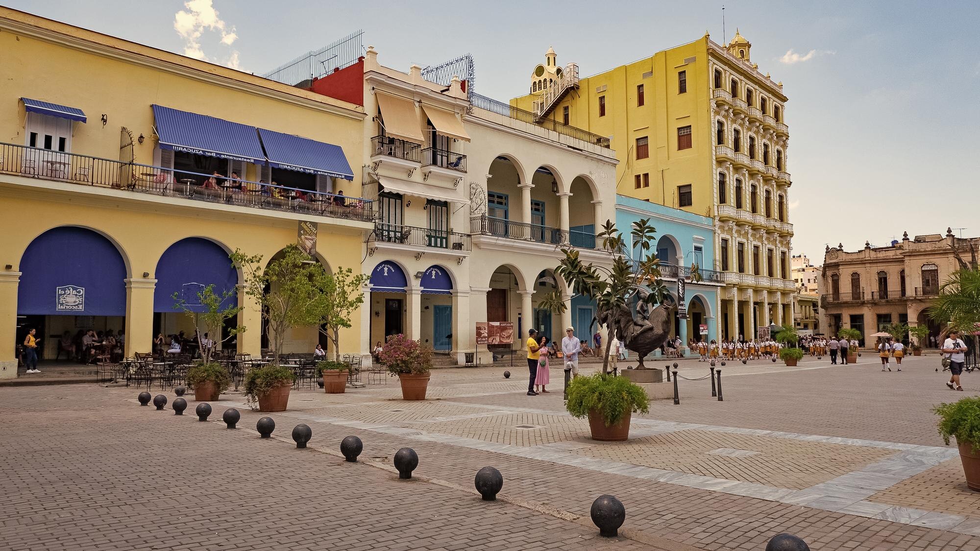 People stroll across a cobbled Old Havana square lined with colorful colonial buildings and potted trees.