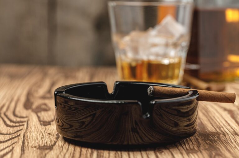 An unbanded cigar rests in a black ashtray on a wooden table, with a glass of amber liquid behind it.